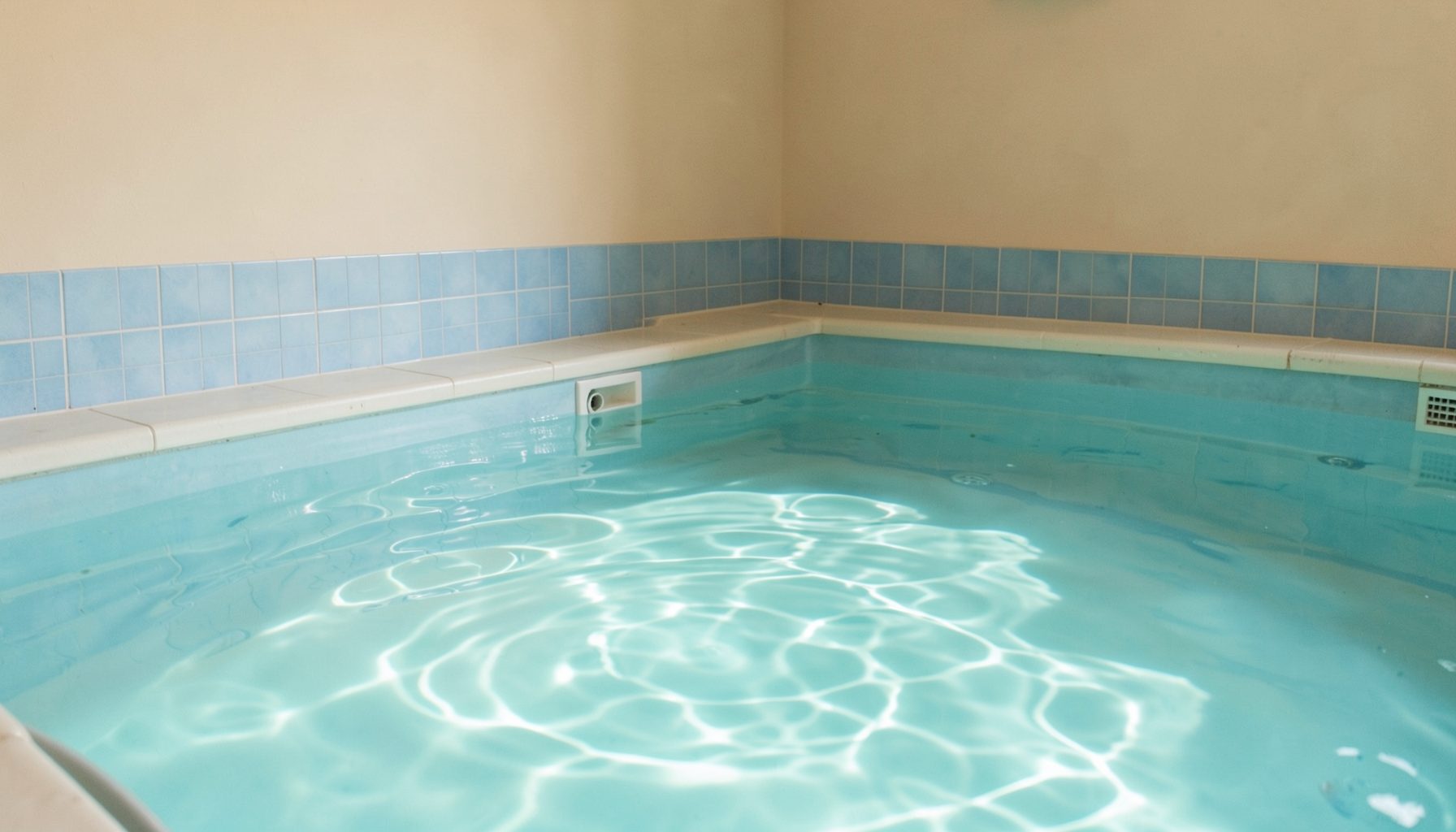 A calm indoor children's pool with soft turquoise water, gentle ripples catching light, surrounded by warm beige walls